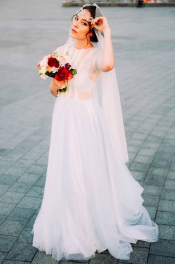Beyaz duvak ve gelin buketi açık havada günbatımında poz tutan postiş güzel Doğu tarzı gelinle