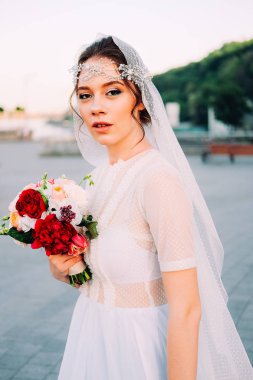 Beyaz duvak ve gelin buketi açık havada günbatımında poz tutan postiş güzel Doğu tarzı gelinle