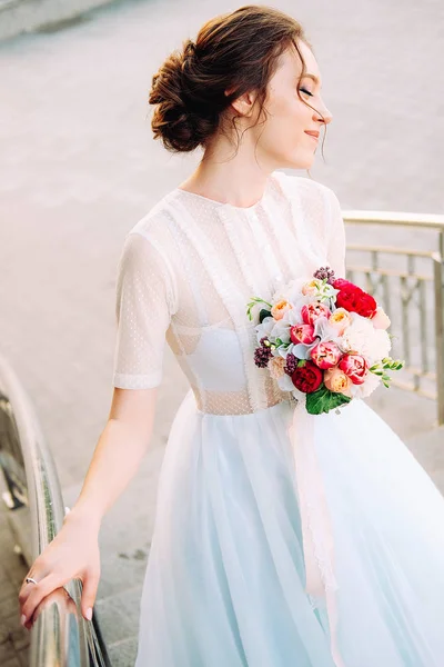 可爱的年轻新娘与新娘花束制成的牡丹 小苍兰花和穿着白色薄纱婚纱站在楼梯上 — 图库照片