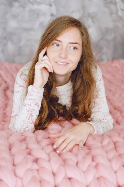 Young woman sitting at the bed on warm peach color throw blanket smiling and looking happy