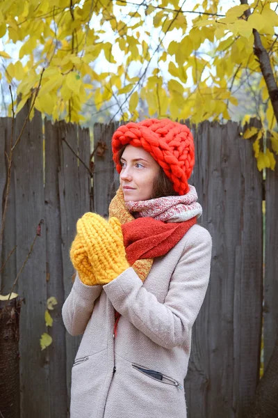 Colorful fall scenery. Young woman in beige wool coat and large knitted red hat and yellow mittens walking outdoorin countryside.