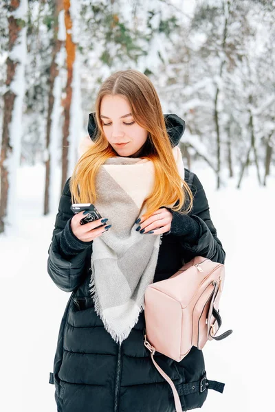 迷人的金发碧眼的年轻成年妇女走在冬季森林充满了雪韦林格休闲服装黑色帕卡 牛仔裤 粉红色皮靴和背包和米色披肩和发短信给朋友 — 图库照片