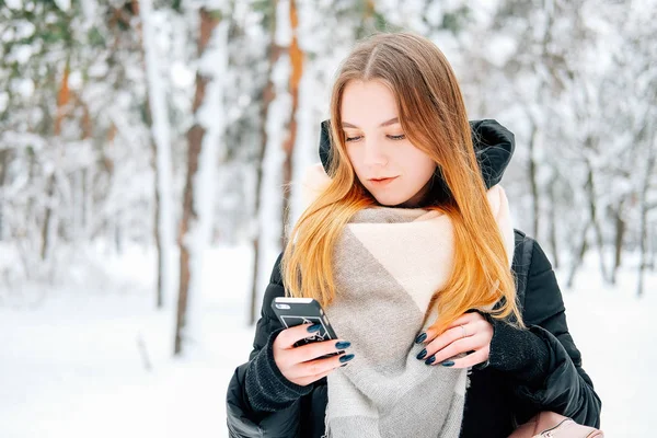 迷人的金发碧眼的年轻成年妇女走在冬季森林充满了雪韦林格休闲服装黑色帕卡 牛仔裤 粉红色皮靴和背包和米色披肩和发短信给朋友 — 图库照片