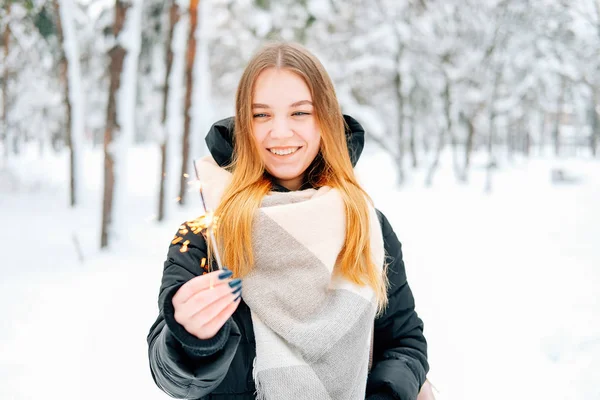 迷人的金发碧眼的年轻成年妇女走在冬季森林充满雪穿着休闲服装的黑色帕卡 牛仔裤 粉红色皮靴和背包和米色披肩 并举行烟花火花 — 图库照片