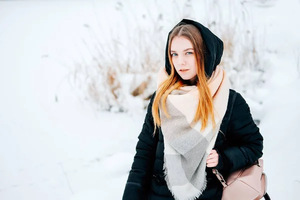 冬の森を歩いて魅力的な金髪の若い成人女性雪の黒のパーカー ジーンズ ピンクの革ブーツとバックパックとベージュのショールのカジュアルな服を身に着けています — ストック写真
