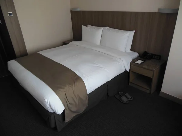 Otel yatak odasında kahverengi ve beyaz renkler: kral yatak, temiz beyaz çarşaf, kahverengi başucundaki ile telefon ve Not defteri, terlik katta