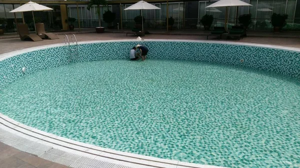 Round swimming pool in hotel with chairs around it, no water inside and maintenance workers trying to fix problem