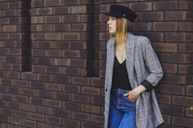 Genç kız güzel bir ceket, kot pantolon ve bir tuğla duvar arka plan üzerine şapka