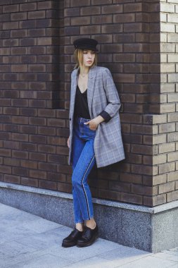 Genç kız güzel bir ceket, kot pantolon ve bir tuğla duvar arka plan üzerine şapka