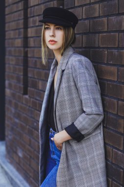 Genç kız güzel bir ceket, kot pantolon ve bir tuğla duvar arka plan üzerine şapka