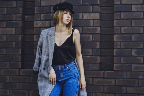 Genç kız güzel bir ceket, kot pantolon ve bir tuğla duvar arka plan üzerine şapka
