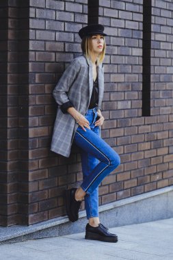 Genç kız güzel bir ceket, kot pantolon ve bir tuğla duvar arka plan üzerine şapka