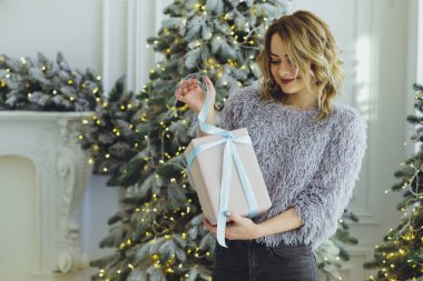 Yeni yıl hediyeleri ile Noel ağacı yakınındaki ellerinde bir hediye ile gri süveterli genç güzel sarışın kız