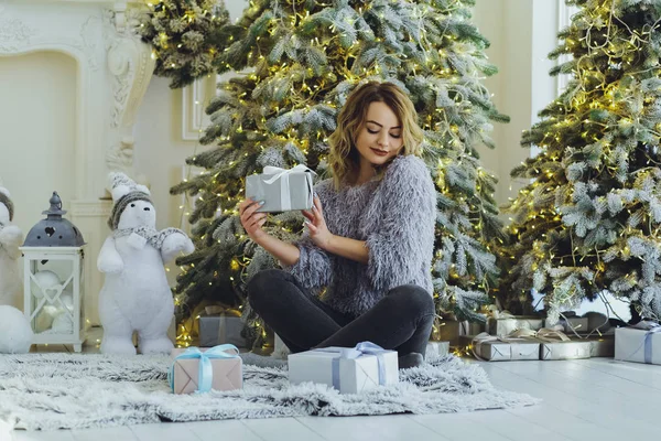 Yeni yıl hediyeleri ile Noel ağacı yakınındaki ellerinde bir hediye ile gri süveterli genç güzel sarışın kız