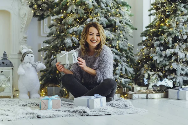 Yeni yıl hediyeleri ile Noel ağacı yakınındaki ellerinde bir hediye ile gri süveterli genç güzel sarışın kız