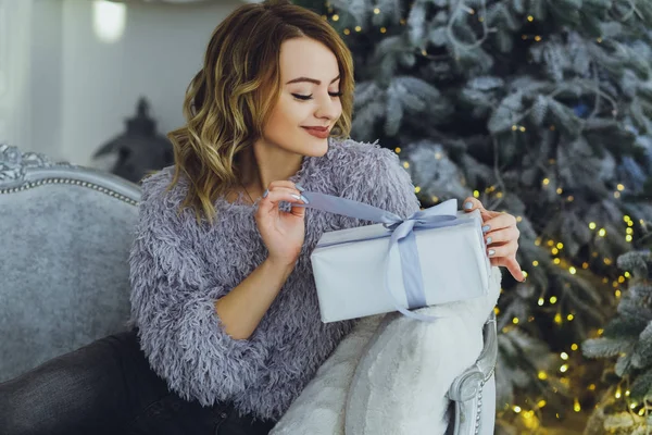 Yeni yıl hediyeleri ile Noel ağacı yakınındaki ellerinde bir hediye ile gri süveterli genç güzel sarışın kadın