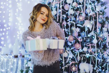 Yeni yıl hediyeleri ile Noel ağacı yakınındaki ellerinde bir hediye ile gri süveterli genç güzel sarışın kadın