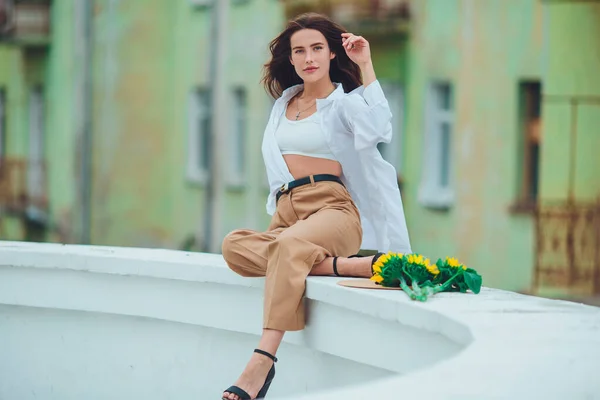 Beautiful young woman with dark hair in a hat, in the city, holding hands for a bouquet of sunflowers and looking away. Fashionable and stylish girl