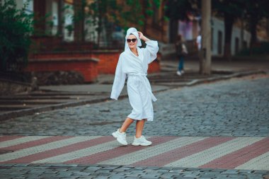 Kafasında beyaz bir havlu ve zarif güneş gözlükleriyle beyaz bornozlu çekici bir kadın büyük bir şehrin caddesinde yürüyor.