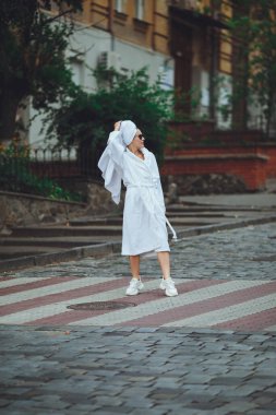 Kafasında beyaz bir havlu ve zarif güneş gözlükleriyle beyaz bornozlu çekici bir kadın büyük bir şehrin caddesinde yürüyor.