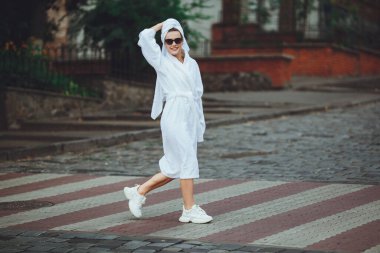 Kafasında beyaz bir havlu ve zarif güneş gözlükleriyle beyaz bornozlu çekici bir kadın büyük bir şehrin caddesinde yürüyor.