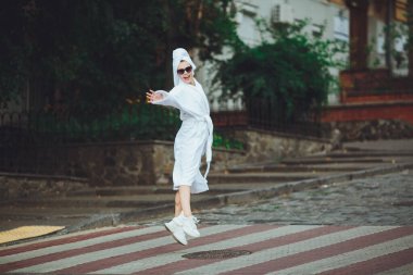 Kafasında beyaz bir havlu ve zarif güneş gözlükleriyle beyaz bornozlu çekici bir kadın büyük bir şehrin caddesinde yürüyor.