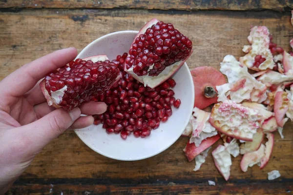 Man holds a pomegranate slice in his hand against the background of a plate with pomegranate fruits in a white plate, near the rind from the pomegranate. — Stock Photo, Image