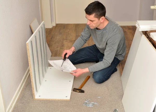 Atractive male assembling a cupboard - reading instructions. — Stock Photo, Image