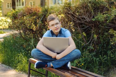 Oğlan uzaktan öğreniyor. Parkta dizüstü bilgisayarı olan bir genç. Mesafesini koruyor. Çocuk bankta tek başına..