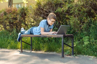 Oğlan uzaktan öğreniyor. Parkta dizüstü bilgisayarı olan bir genç. Mesafesini koruyor. Çocuk bankta tek başına..