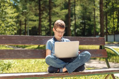 Oğlan uzaktan öğreniyor. Parkta dizüstü bilgisayarı olan bir genç. Mesafesini koruyor. Çocuk bankta tek başına..