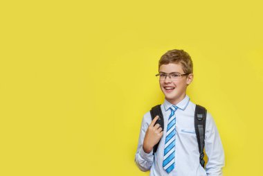 Okul üniformalı, sırt çantalı, sarı arka planda gülümseyen bir çocuk..