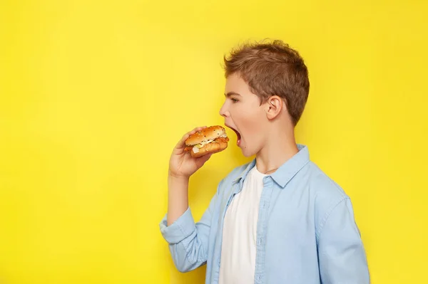 A teenager bites a burger on a yellow background in profile. A side view.