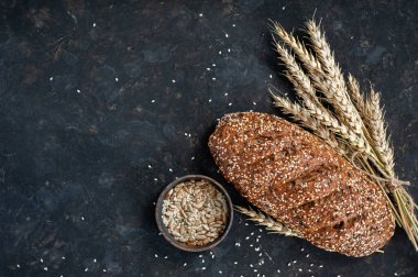 Buğday kulakları, tohumlar ve susam tohumlarıyla koyu bir zemin üzerinde taze kepekli ekmek..