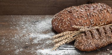 Kahverengi ahşap zemin üzerinde iki farklı somun kepekli ekmek un, tohum, susam tohumu ve buğday kulağı..