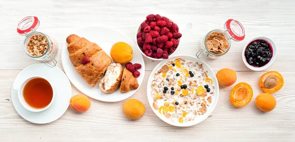 Ahududu ve kayısı ile yaz kahvaltısı. Süt ve yaban mersinli granola. Beyaz kaplarda yağlı reçel ve kruvasan. Bir fincan çay ve bir bardak meyve suyu beton gri-mavi arka planda.