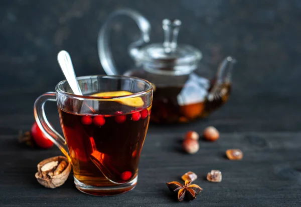 Herbstlicher heißer Tee in einer Glasschale mit Beeren, Äpfeln und Nüssen auf schwarzem Holzgrund. Ein wärmendes Getränk. — Stockfoto