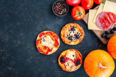 Cadılar Bayramı 'nda örümcekler ve hayaletlerle süslenmiş mini pizzalar. Koyu arkaplanda malzemeler ve balkabakları olan bir kompozisyon. Çocuklar için Cadılar Bayramı yemeği. Metin için yer.