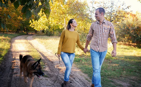 Couple d'amour heureux marchant dans le parc avec un chien de berger allemand, se tenant la main et se regardant mutuellement — Photo