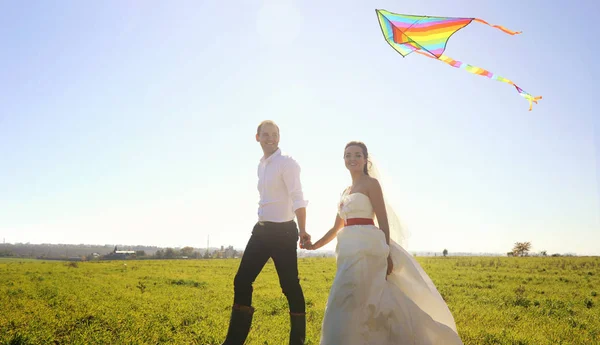 Happy wedding couple walking on green field with flying kite — Stock Photo, Image