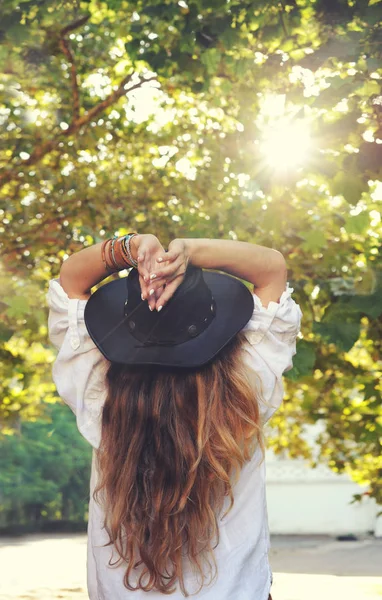 Молодая женщина в стиле фо наслаждается солнечным светом в летнем парке, хиппи, инди-стиль, шикарная кожаная черная шляпа, длинные волосы, концепция путешествия, вид сзади — стоковое фото