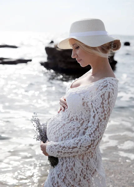 Schwangere am Meer in weißem Spitzenkleid und Hut. — Stockfoto