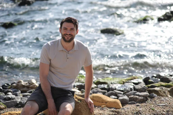 Junger stylischer Mann im Freien, weißes Poloshirt, sitzt auf einem Stein — Stockfoto