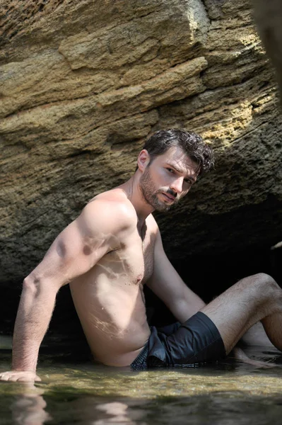 Jovem homem topless perto das rochas do mar sentado na água e olhando para a câmera — Fotografia de Stock
