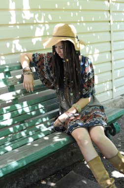 Boho tarzı elbisesi ve şapkası olan güzel bir kadın evinin yanında bankta oturuyor.