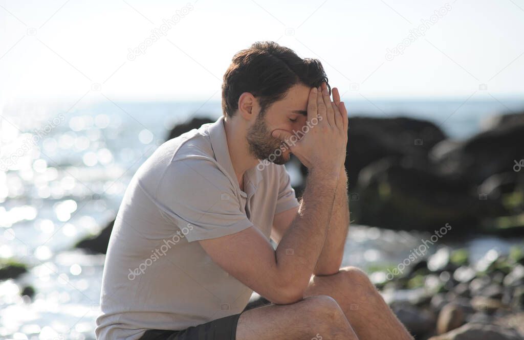 Depressed sad young man sitting on a sea beach, palms on face
