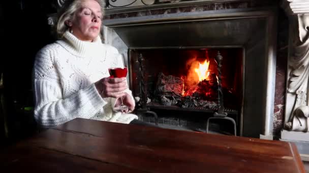 单身老妇人穿着白色毛衣在燃烧的壁炉前喝红酒 孤独的老妇人喝红酒在燃烧的壁炉前老年妇女喝红酒在家里喝红酒 — 图库视频影像