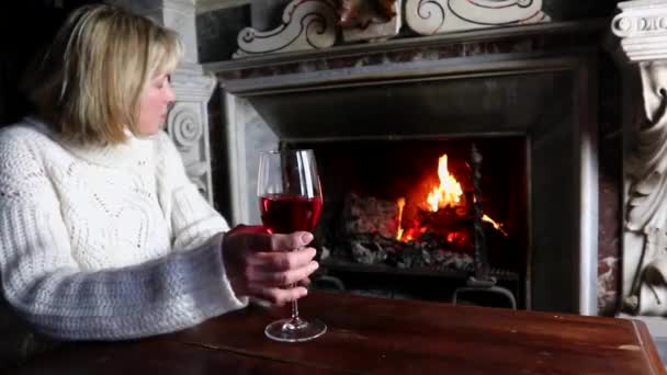 单身老妇人穿着白色毛衣在燃烧的壁炉前喝红酒 孤独的老妇人喝红酒在燃烧的壁炉前老年妇女喝红酒在家里喝红酒 — 图库视频影像