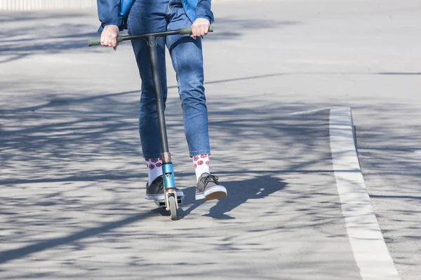 Las piernas de una chica desconocida en zapatillas blancas y jeans ajustados montan en un scooter eléctrico negro sobre un asfalto urbano oscuro — Foto de Stock