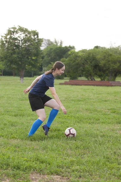 Young female soccer or football player with long hair in sportwear and boots kicking ball for the goal in jump at the stadium.